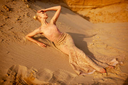 Fashionable blonde girl model in gold dress, touching her face with hands, have closed eyes, lying on a warm sand.の写真素材