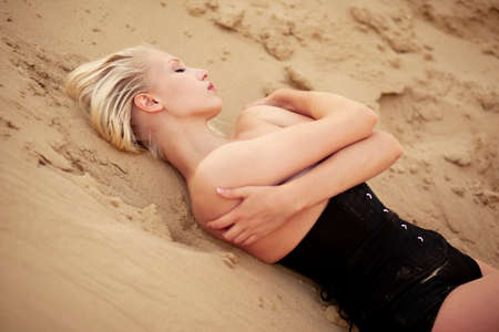 Sexy blonde girl model in black corset with crossed hands, have closed eyes, lying on a warm sand. Fashionable model.の写真素材