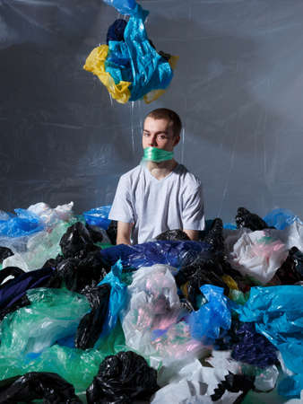 Young man sitting down, tied to the mouth with a plastic bag, surrounded by plastic bags, looking at...