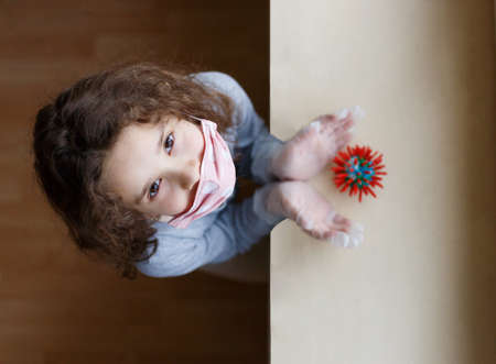 A little girl wears a mask and medical gloves, looks up at the camera, holding her hands around a symbol of Covid-19 virus. Danger of infection, protection needed to avoid infection.の写真素材