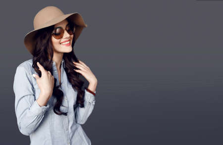Cheerful model with round sunglasses, curly hair, wearing in jeans shirt and beige hat, touches his brown hair.の写真素材