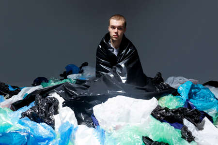 Confident young man with haircut seated on plastic bag garbage, environmental - consciousness concept, shoot inside in studio, dark light background.の写真素材