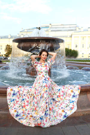 Beautiful young woman with red hair dressed in long dress sitting near fountain in Moscow city.の写真素材