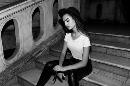 Fashion portrait of a fashion girl in stylish hat, with straight hair, looking down seated on stairs in the night street.の写真素材