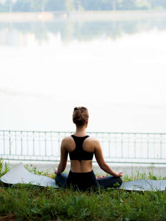 Back of a young attractive woman practicing yoga, sitting in Lotus pose, wearing in sportswear in the park.の写真素材