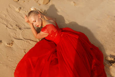 Graceful blonde girl in a red dress lies on the sand. Fashionable female model posing on sand in beautiful red dress.の写真素材