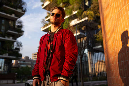 Portrait of the young man with sunglasses and a red jacket, looking confident in the city being sunset.の写真素材