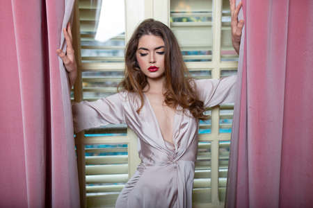 Seductive young woman with long waving hair and makeup posing in silk pink dress near big windows with curtains.の写真素材