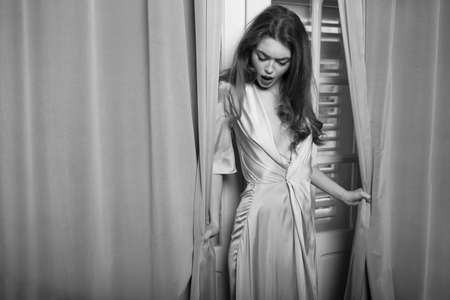 Seductive young woman with long waving hair and makeup posing in silk pink dress near windows with curtains.の写真素材