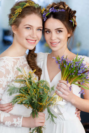 Vertical portrait of two brides in white wedding dresses with flowers in their hair, with a bouquet of wild flowers.の写真素材