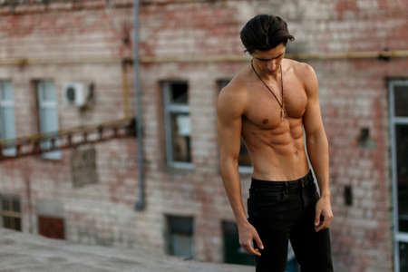 Athlete strong young man shirtless dressed in black jeans, looking down posing with attitude on old roof outside.の写真素材