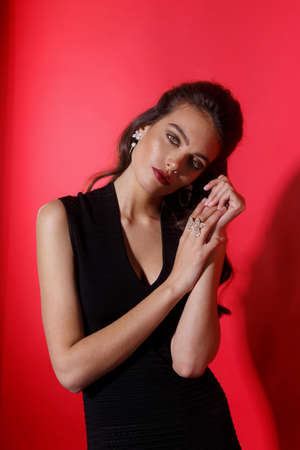 Sensual woman in black dress with hairstyle, holding hands crossed with luxury jewellery, over red...