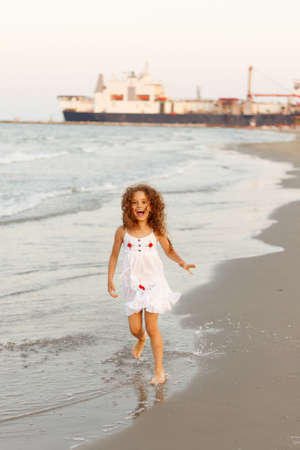 Little curly hair girl jumping on the beach with a white dress, seascape and massive ship backgroundの写真素材