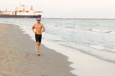 Runner training outside working at the beach. Fit young male sport fitness model exercising in full body in summer.の写真素材