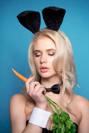 Portrait of a sexy blonde woman with makeup, bunny ears, holding a carrot in her hand, over blue background.の写真素材