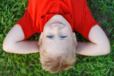 Close up portrait of cute smiling little boy with blue eyes , wear red shirt lying on grass in park looking at camera.の写真素材
