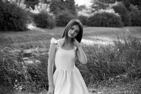 Frontal portrait of a beautiful young girl dressed in dress seated on grass, touching her neck, looking at camera.の写真素材