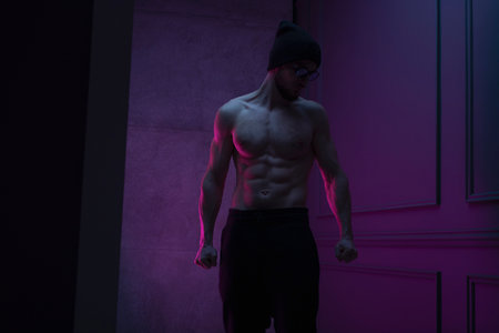 Profile portrait of a shirtless bearded young man in hat posing in studio with concrete wall with purple neon color.の写真素材