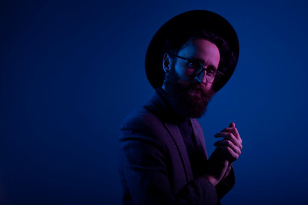 Portrait of a bearded man in hat and eyeglasses looking at camera, isolated on dark neon purple color background.の写真素材