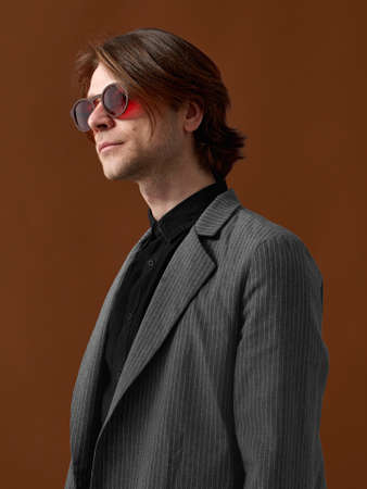 Attractive elegant businessman in red eyeglasses, standing with a firm posture on brown studio...