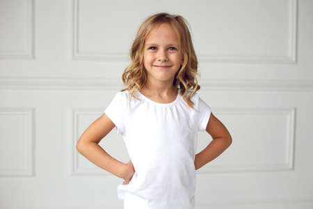 Little girl smiling and looking at the camera, in a white T-shirt, holding hands on her waist, on a white background.の写真素材