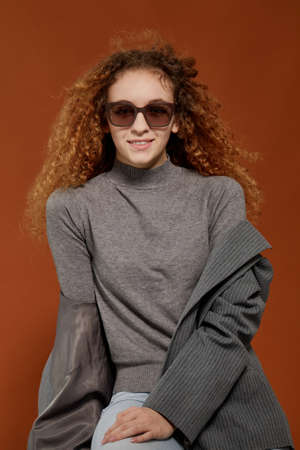 Curly girl with attitude, wearing grey clothes and sunglasses looking at the camera on brown...
