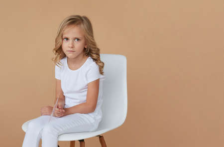 Closeup front portrait of little girl with wavy blond hair, dressed in white, sitting on chair, over beige background.の写真素材