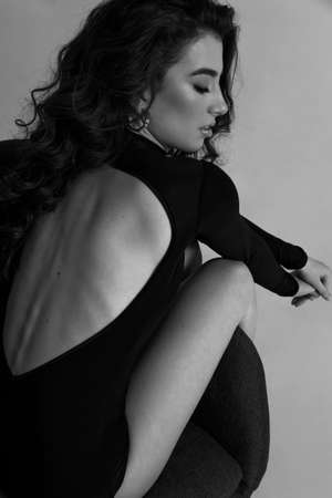 Back view of a sensual young woman in a black bodysuit sitting on chair, on a grey background. Vertical view.の写真素材