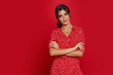 Portrait of a sensual young woman with cross hands, wearing red dress, makeup, ponytail, isolated on a red background.の写真素材