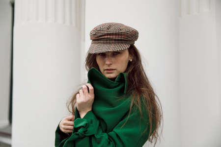 Closeup front portrait of a stylish young woman with curly hair, wearing green coat and hat, columns background.の写真素材