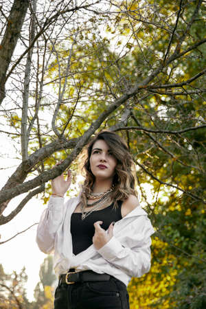 Portrait of a young woman in white shirt and black jeans near green tree with sunset light and sunbeams.の写真素材