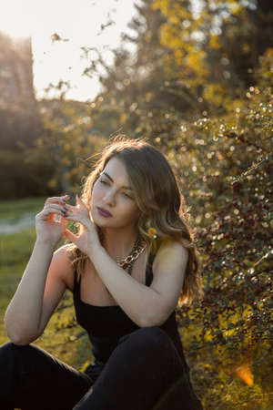 Sensual pensively young woman with makeup and curly hair sitting down on grass in the park with sunset light and trees.の写真素材