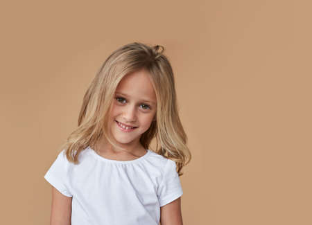 Closeup front of a pretty little girl with wavy blond hair, dressed in white clothes, over beige background.の写真素材