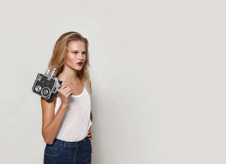 Young, stylish blonde girl standing in a profile holding on a shoulder a retro camera, over white background.の写真素材