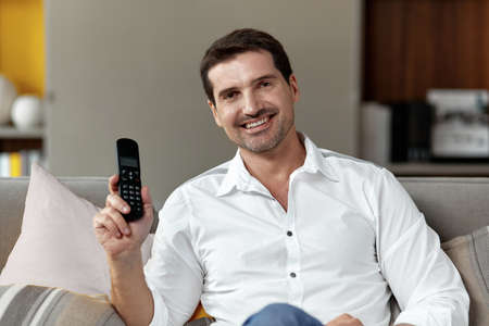 Front image of a handsome smiling man looking at cameraa holding in hands a stationary phone sitting on a couch.の写真素材