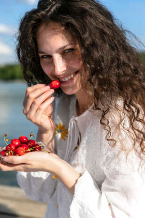 Close up photo of cheerful cute nice sweet curly girl with lips pomaded holding a cherry with hand, outside portrait.の写真素材