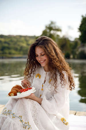 Happy woman with curly hair enjoy picnic by the lake outdoors, eat croissant. Delicious and healthy picnic in nature.の写真素材