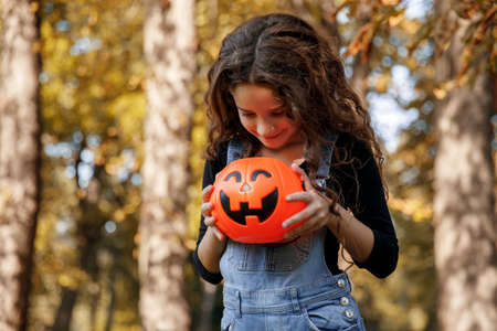 A pretty little girl with curly hair, holding a halloween orange cauldron in her hand, on a sunny autumn day background.の写真素材