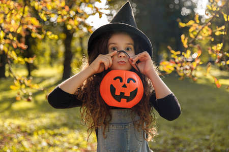 Cute little curly girl with curly hair in black witch hat making faces, holding a halloween orange cauldron in her hand, on a sunny autumn day background.の写真素材