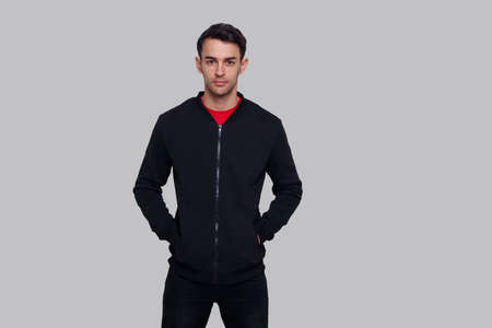 Frontal image of a smiling young brunette man in dark blue jumper and jeans, isolated over grey...
