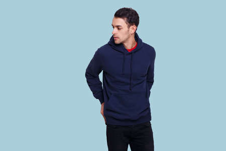 Profile of a young man in dark blue jumper and jeans, holding his hands in his pockets, isolated light blue background.の写真素材