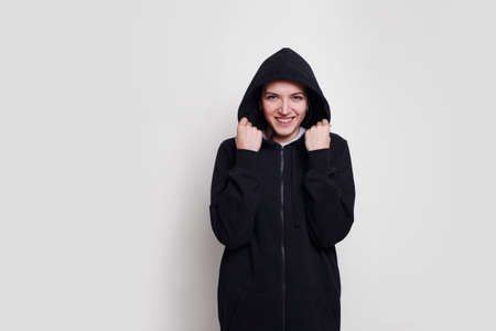 Frontal portrait of a happy brunette girl in a black sweater with hood on her head, isolated on white background.の写真素材