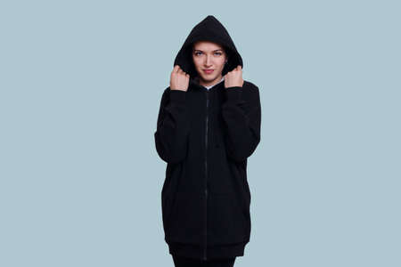 Frontal portrait of a brunette girl dressed in a black sweater with hood on her head, isolated on blue background.の写真素材