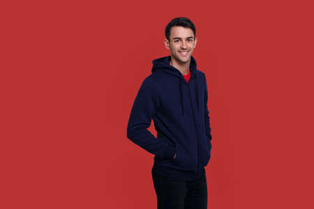 Happy handsome young brunette man in dark blue jumper and jeans over red background. Copy space.の写真素材