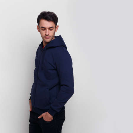 Young man in dark blue jumper and jeans, with hands in his pocket, looking down, isolated white background.の写真素材