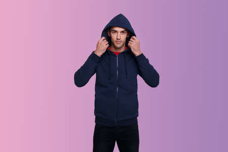 Portrait of a confident young man in dark blue jumper and jeans, with a hood on his head, isolated purple background.の写真素材