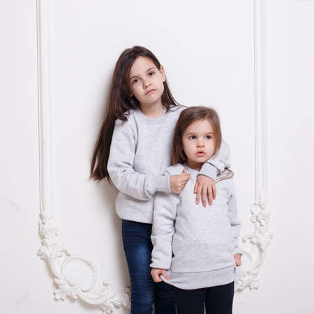 Two adorabile littles girl in sweater and jeans, posing together embraced on white background.の写真素材