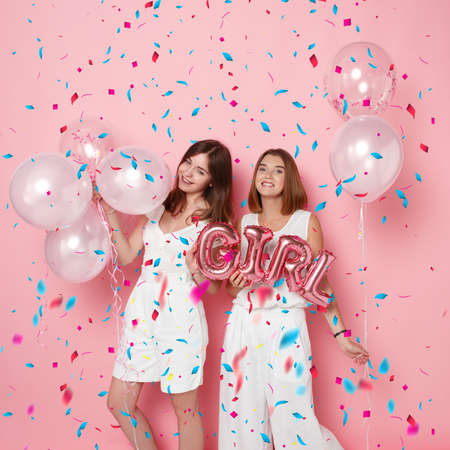 Portrait of happy young women, dressed in a white, holdings a balloons, over colored confetti, pink background.の写真素材