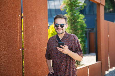 Young hipster man in sunglasses stand near building wall outdoors, holding a mobile cell phone and smiling.の写真素材