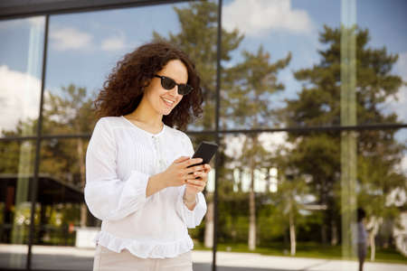 Close-up portrait of a girl with curly hair, looking at the cell phone, against a building with window background.の写真素材
